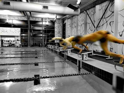 A swimming pool with lanes marked, embedded within an indoor facility with large windows and wall decorations. Multiple swimmers are in mid-air, diving off starting blocks into the pool.