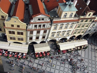 An aerial view captures a picturesque European street lined with historic buildings, showcasing their distinct architectural features such as red-tiled roofs and ornate facades. Below, a bustling square is populated with people, and several outdoor cafes with umbrellas and seating arrangements, suggesting a lively atmosphere.