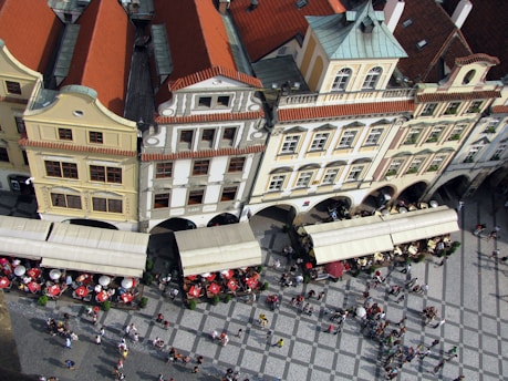 An aerial view captures a picturesque European street lined with historic buildings, showcasing their distinct architectural features such as red-tiled roofs and ornate facades. Below, a bustling square is populated with people, and several outdoor cafes with umbrellas and seating arrangements, suggesting a lively atmosphere.