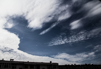 The image features a clear sky with dramatic cloud formations. The clouds range from thin, wispy streaks to thicker, fluffier sections, with a darker, almost stormy appearance towards the top. The lower portion of the image shows the roofline of a building with some visible chimneys and vents.