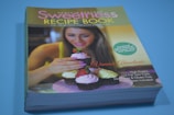 A recipe book titled 'Sweetness Recipe Book' is displayed with a cover image of a woman decorating cupcakes with pink frosting and a strawberry. The book highlights over 100 sweet protein-packed recipes. There is a starburst text referring to calorie information, and a list of nutrition-related claims.