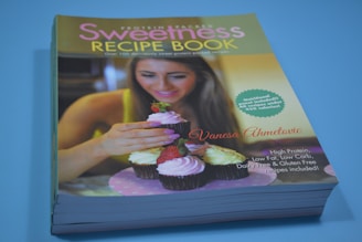 A recipe book titled 'Sweetness Recipe Book' is displayed with a cover image of a woman decorating cupcakes with pink frosting and a strawberry. The book highlights over 100 sweet protein-packed recipes. There is a starburst text referring to calorie information, and a list of nutrition-related claims.