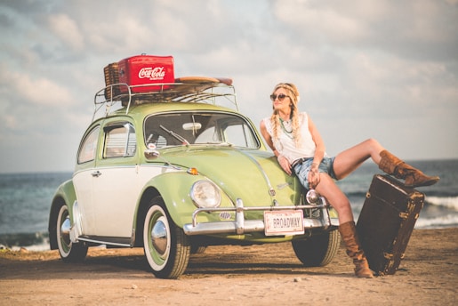 A vintage green Volkswagen Beetle is parked on a sandy beach. A woman wearing a white top, denim shorts, sunglasses, and brown boots leans casually against the car. The car's roof rack holds a wicker basket and a red Coca-Cola cooler. The trunk is open, showcasing a traveler's suitcase. The setting gives off a retro vibe with a backdrop of the ocean and a cloudy sky.