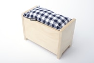 A rectangular wooden box with a simple, minimalist design features a cushion on top. The cushion has a black and white checkered pattern and a small tag with the name 'LILIANE' attached to it. The box is light-colored wood with clean, smooth edges.