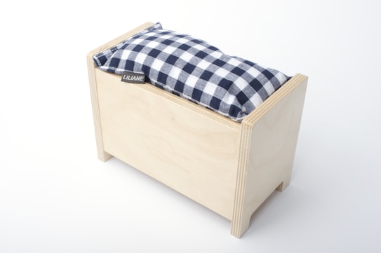 A rectangular wooden box with a simple, minimalist design features a cushion on top. The cushion has a black and white checkered pattern and a small tag with the name 'LILIANE' attached to it. The box is light-colored wood with clean, smooth edges.
