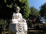 A serene white stone statue of a seated figure in a meditation pose, with one hand raised in a traditional gesture. The statue is surrounded by lush green trees in a garden setting. Sunlight filters through the foliage, casting gentle shadows on the ground. A building with a red roof can be seen in the background.
