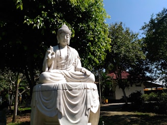 A serene white stone statue of a seated figure in a meditation pose, with one hand raised in a traditional gesture. The statue is surrounded by lush green trees in a garden setting. Sunlight filters through the foliage, casting gentle shadows on the ground. A building with a red roof can be seen in the background.