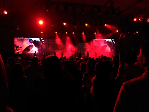 The image captures a lively concert scene with an enthusiastic crowd. The stage is illuminated with vibrant red lighting and smoke effects. A large screen displays visuals, possibly related to the performance. Silhouettes of people raising their hands can be seen against the bright lights.