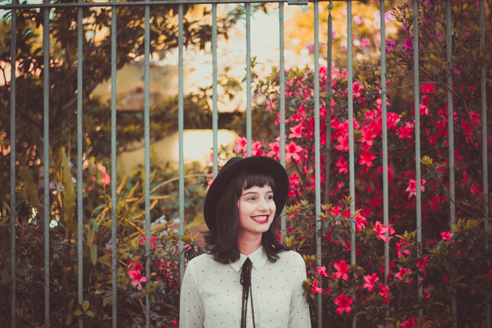 A young woman in a hat smiling while standing next to flower bushes