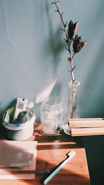 A wooden table holds various objects including a toothbrush, a sheet of paper, a small dish with steel wool and a tube, a transparent jar, a tall glass vase with a dried branch, and several wooden pieces. The background is a plain wall, and sunlight casts shadows across the surface.
