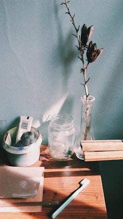 A wooden table holds various objects including a toothbrush, a sheet of paper, a small dish with steel wool and a tube, a transparent jar, a tall glass vase with a dried branch, and several wooden pieces. The background is a plain wall, and sunlight casts shadows across the surface.