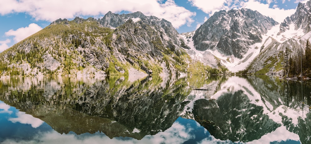 rocky mountains covered with green grass reflected on body of water under white and blue cloudy sky
