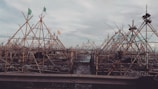 A complex structure of bamboo scaffolding is assembled against a cloudy sky. Flags are visible atop several of the triangular frames, and the wooden slats create an intricate pattern.