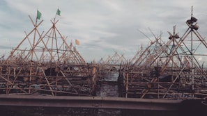 A complex structure of bamboo scaffolding is assembled against a cloudy sky. Flags are visible atop several of the triangular frames, and the wooden slats create an intricate pattern.