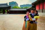 A family dressed in traditional Korean hanboks stands in front of a historic palace, with modern buildings visible in the background. The setting combines historical architecture with urban elements.