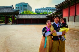 A family dressed in traditional Korean hanboks stands in front of a historic palace, with modern buildings visible in the background. The setting combines historical architecture with urban elements.