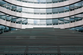 A modern architectural facade featuring a symmetrical pattern of horizontal lines and reflective glass windows. The building's surface is composed of metallic panels and glass, creating a sleek and contemporary appearance.