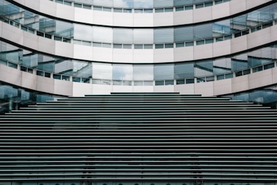 A modern architectural facade featuring a symmetrical pattern of horizontal lines and reflective glass windows. The building's surface is composed of metallic panels and glass, creating a sleek and contemporary appearance.