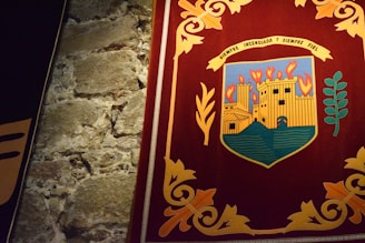 A richly colored tapestry depicting an emblem or coat of arms featuring a castle with flames above it, signifying burning. The emblem is set against a dark red background bordered by intricate golden floral patterns. Adjacent is a texture of rough, aged stone wall.