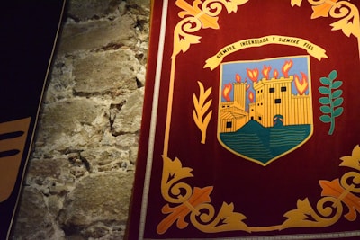 A richly colored tapestry depicting an emblem or coat of arms featuring a castle with flames above it, signifying burning. The emblem is set against a dark red background bordered by intricate golden floral patterns. Adjacent is a texture of rough, aged stone wall.