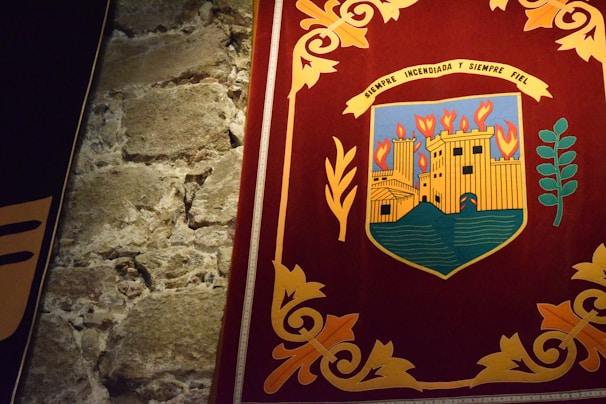 A richly colored tapestry depicting an emblem or coat of arms featuring a castle with flames above it, signifying burning. The emblem is set against a dark red background bordered by intricate golden floral patterns. Adjacent is a texture of rough, aged stone wall.