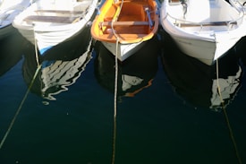 A group of small boats tethered together, floating on calm water. The boats have a clean, white exterior with one featuring an orange interior. Their reflections are visible in the deep, dark blue water, adding a sense of tranquility.