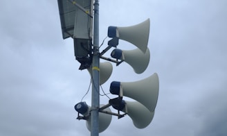 A tall pole with multiple large loudspeakers attached, pointing in various directions during an overcast day. A small box, possibly a control unit, is mounted at the top alongside a solar panel.