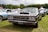 A classic black muscle car is prominently displayed at an outdoor car show. The car is surrounded by other vintage vehicles, and there are tents and people in the background. The setting appears to be a grassy field with trees in the distance under a partly cloudy sky.