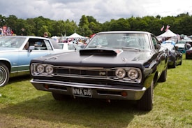 A classic black muscle car is prominently displayed at an outdoor car show. The car is surrounded by other vintage vehicles, and there are tents and people in the background. The setting appears to be a grassy field with trees in the distance under a partly cloudy sky.