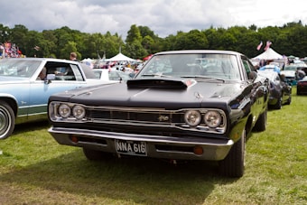 A classic black muscle car is prominently displayed at an outdoor car show. The car is surrounded by other vintage vehicles, and there are tents and people in the background. The setting appears to be a grassy field with trees in the distance under a partly cloudy sky.