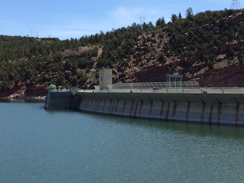 A large dam structure spans across a body of calm water, with the dam itself featuring industrial elements like metal railings and a control tower. The surrounding area consists of rugged terrain with dense greenery and pine trees covering the hills in the background. Overhead, the sky is clear and blue, with visible electrical pylons atop the hills.