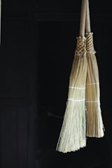 Two traditional straw brooms hang side by side against a dark background. The brooms are bound with string and have a natural beige color, with the wooden handles extending upwards.