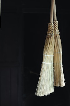 Two traditional straw brooms hang side by side against a dark background. The brooms are bound with string and have a natural beige color, with the wooden handles extending upwards.