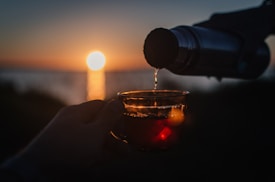 A thermos is pouring a warm beverage into a clear glass cup held by a hand. The scene is set against a backdrop of a sunset over a body of water, with the sun casting golden reflections on the surface.