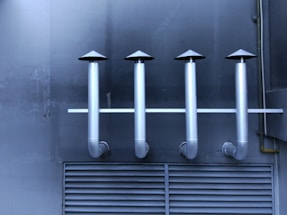 Four metallic ventilation pipes with cone-shaped tops are aligned horizontally against a dark industrial wall. Below the pipes, a vented panel is visible, contributing to the industrial aesthetic.