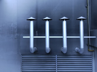 Four metallic ventilation pipes with cone-shaped tops are aligned horizontally against a dark industrial wall. Below the pipes, a vented panel is visible, contributing to the industrial aesthetic.