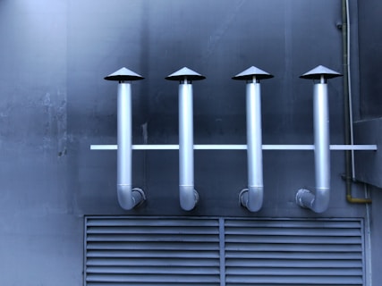 Four metallic ventilation pipes with cone-shaped tops are aligned horizontally against a dark industrial wall. Below the pipes, a vented panel is visible, contributing to the industrial aesthetic.
