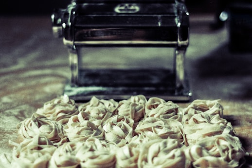 A collection of freshly made pasta nests is placed on a floured surface in front of a pasta machine. The setting appears to be a kitchen, with the focus on the raw pasta ready for cooking.