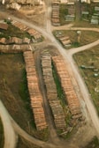 Aerial view of a lumber yard with neatly stacked rows of wooden logs on dirt roads, surrounded by patches of grass and open land. The logs are arranged in long, parallel lines beside intersecting pathways.