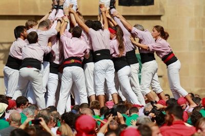 A group of people dressed in white pants and pink shirts with black waistbands are forming a human tower. They are supporting each other by holding onto one another's shoulders and backs. A large crowd is gathered around them, wearing various colors such as red and green.