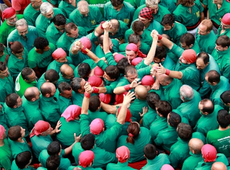 A large group of people dressed in green shirts and red headscarves are gathered closely together, with some individuals raising their arms. The atmosphere appears lively and collaborative, as if they are participating in a collective event or ceremony.