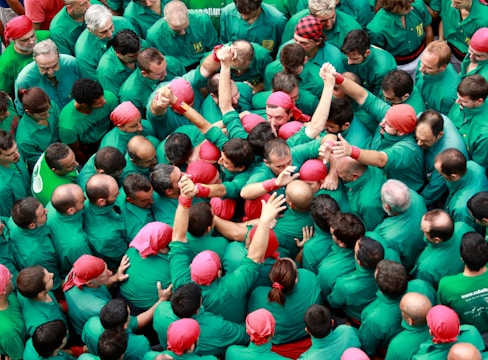 A large group of people dressed in green shirts and red headscarves are gathered closely together, with some individuals raising their arms. The atmosphere appears lively and collaborative, as if they are participating in a collective event or ceremony.