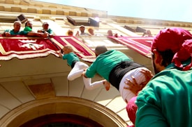 People are participating in a human tower event. Participants in green shirts are forming the tower beneath a balcony decorated with red and white banners. A child is climbing upward to reach the top, assisted by others.