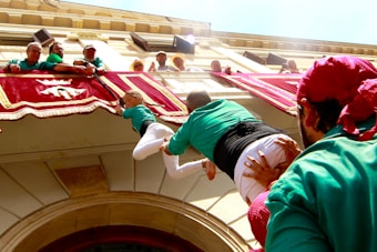 People are participating in a human tower event. Participants in green shirts are forming the tower beneath a balcony decorated with red and white banners. A child is climbing upward to reach the top, assisted by others.