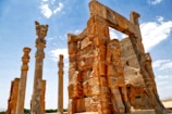 Ancient ruins featuring tall, ornate columns and large stone carvings set against a blue sky. The architecture includes intricate stone reliefs depicting figures and animals, possibly from a historical or archaeological site.
