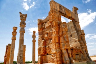 Ancient ruins featuring tall, ornate columns and large stone carvings set against a blue sky. The architecture includes intricate stone reliefs depicting figures and animals, possibly from a historical or archaeological site.