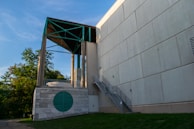 A modern building with a large concrete facade and a unique architectural design. It features a stairway leading to a door, a rooftop structure with teal accents, and a circular vent on a marble wall. Surrounding the building are green trees and a small patch of grass beneath a clear blue sky.