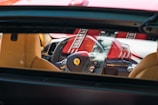 The interior of a luxury car is visible through the rear window, highlighting the steering wheel with a prominent logo, beige leather seats, and a red and black dashboard. Sunlight creates bright reflections on the surfaces.