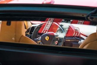 The interior of a luxury car is visible through the rear window, highlighting the steering wheel with a prominent logo, beige leather seats, and a red and black dashboard. Sunlight creates bright reflections on the surfaces.