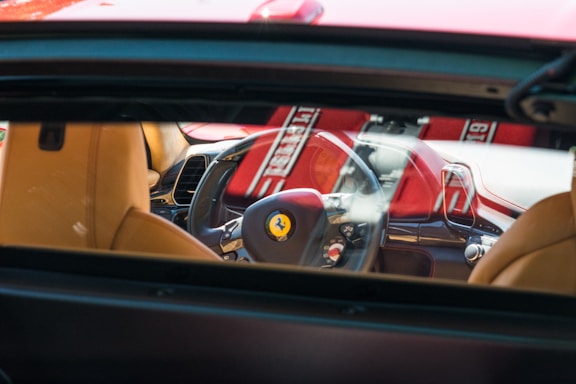 The interior of a luxury car is visible through the rear window, highlighting the steering wheel with a prominent logo, beige leather seats, and a red and black dashboard. Sunlight creates bright reflections on the surfaces.